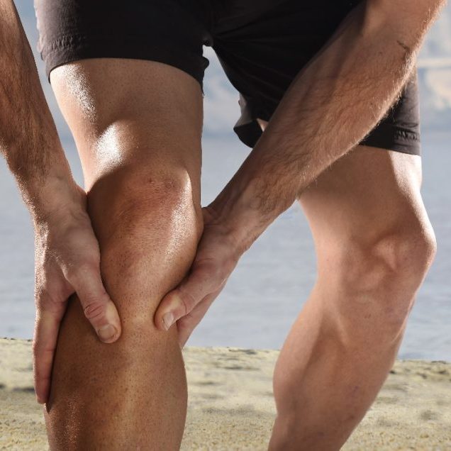 young sport man with strong athletic legs holding knee with his hands in pain after suffering muscle injury during a running workout beach training in muscular or ligament wound
